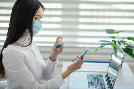 Disinfecting. Young woman in a protective mask spraying disinfector on her smartphone
