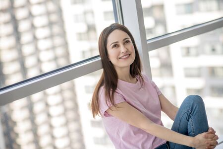 Great mood. Beautiful dark-haired woman in a pink tshirt and jeans sitting by the window, looking...