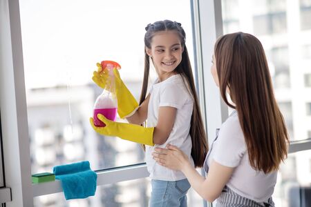 Time for cleaning. Smiling daughter in rubber gloves with a detergent turned to her mom standing behind, both near the window.の写真素材