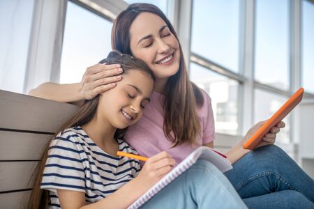 Happy people. Happy young mother with her daughter of primary school age sitting on the floor close to each other, smiling, closing her eyes.の写真素材