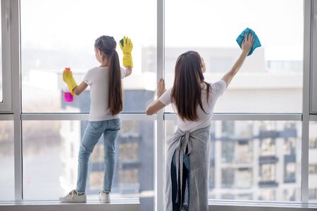 Clean windows. Young mother with her teenage daughter washing together a large window at home, standing with their backs.の写真素材