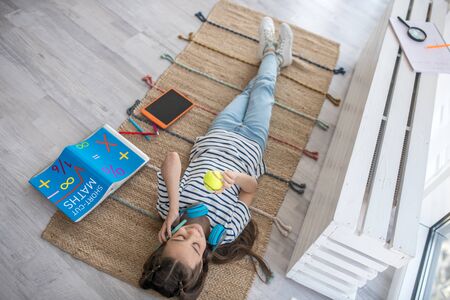 Pastime at home. Girl with headphones and a small ball talking on a smartphone while lying on rug on the floor near the window of house, having fun.の写真素材