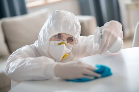 Working hard. Woman in white workwear and protective gloves disinfecting the table surface carefullyの写真素材