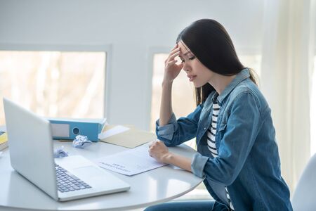 Job loss. Sad dark-haired female sitting at her desk with unemployment claim, touching her nose...