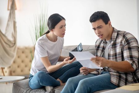 Finance check. Brunette male holding paper, dark-haired female sitting with calcutor, both looking...