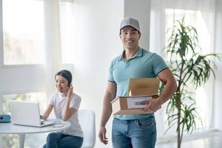 Delivery service. Male courier standing with parcels, dark-haired female typing on laptop behind himの写真素材