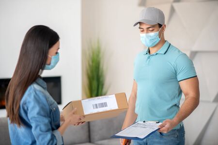 Parcel delivery. Muscular young man with a document, long-haired woman with a parcel, standing opposite each other in protective masks.の写真素材