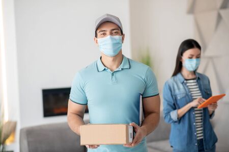 Quarantine, coronavirus. Man in a cap with a box in his hands in the back of the room is a woman with a tablet, both in protective masks.の写真素材