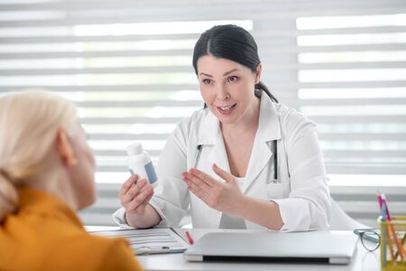 Doctor advice and recommendation. Convincing beautiful woman in a white coat with medicine in her hand telling a blonde patient sitting opposite.の写真素材