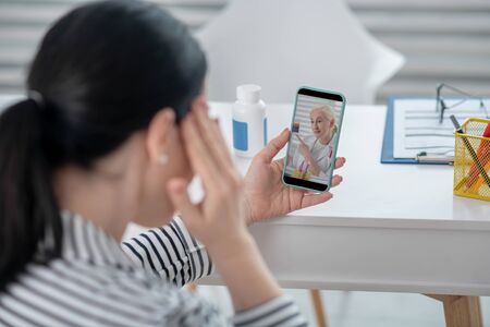 Symptoms, medical consultation. Dark-haired woman looking at a smartphone consulting online, another hand near her head.の写真素材