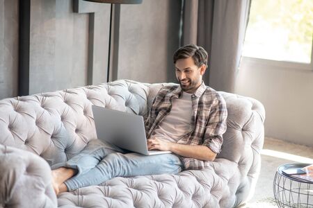 Comfortably at home. Cheerful happy man in jeans lying barefoot on the sofa with a laptop with his back to the window at home.の写真素材