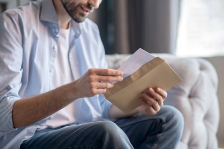 Letter. Hands of a man sitting on a sofa, slowly pulling out a letter from a paper envelopeの写真素材