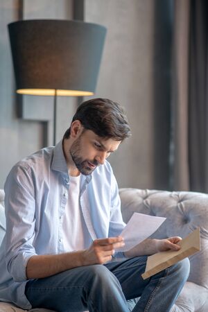 Letter, envelope. Serious young adult man carefully looking at a letter in his hands sitting on a sofa at home.の写真素材