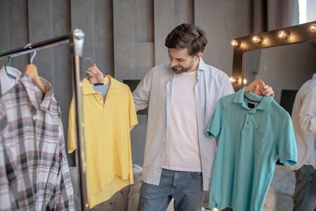 Difficult choice. Bearded dark-haired man standing near the mirror of the house holding in his hands two tshirts on trempels looking at them with interestの写真素材