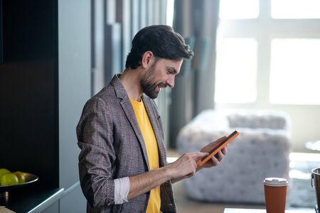 Interesting news. Dark-haired bearded man with a tablet in his hands, at home near the table, looking at the screen smiling in a good moodの写真素材