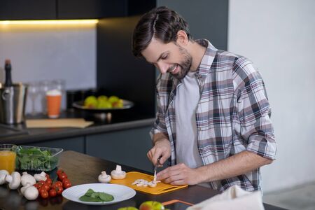 Cooking at home. Young adult man with a beard chopping mushroom champignon, sitting at a table at home, happy.の写真素材