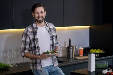 Tasty and healthy food. Smiling young adult man in a white t-shirt and plaid shirt standing in the kitchen with a plate of vegetable salad in his hands.の写真素材