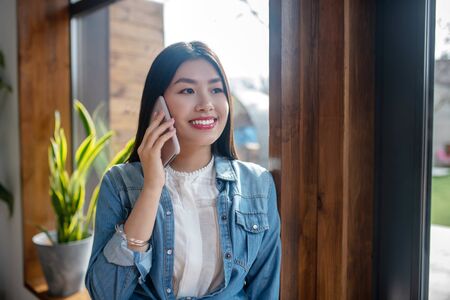 Travel plans. Dark-haired female leaning on door frame, smiling, talking on mobile phone