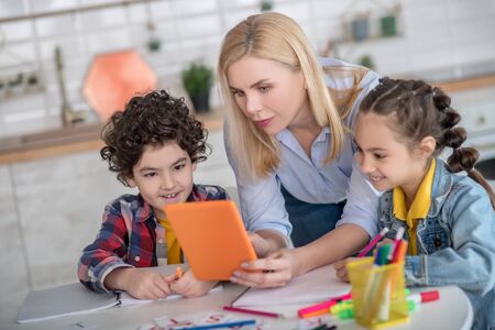 At home. Curly boy and dark-haired girl sitting at table, blonde female bending between them,...