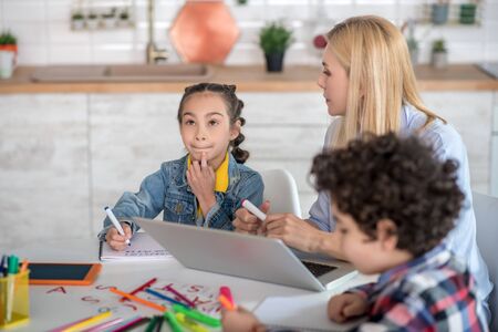 At home. Curly boy and dark-haired girl sitting at table, doing their assignments, blonde female helping them, explaining something to girlの写真素材