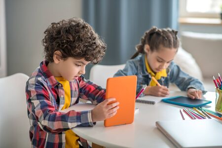 At home. Boy and girl sitting at round table, holding tablets, busy with writingの写真素材