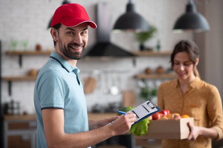 Courrier at work. Young male courrier in a red hat holding groceries and smiling nicelyの写真素材