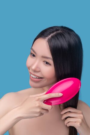 Hair care. Brunette young asian woman brushing her hair and smiling