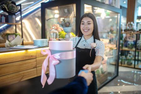 Flower shop. Brunette female in apron giving round gift box with pink bow to customer, receiving credit card from himの写真素材