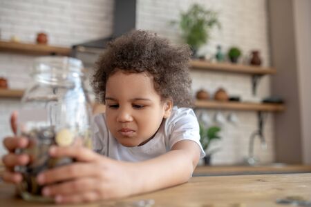 Not much money. Cute curly-haired kid in a white tshirt holding a moneybank and looking unsatisfiedの写真素材
