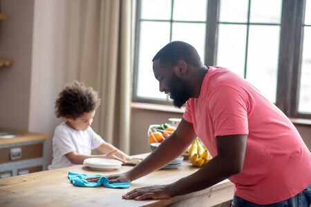 Kitchen cleaning. Tall dark-skinned man in a pink tshirt wiping the table while his kid helpingの写真素材