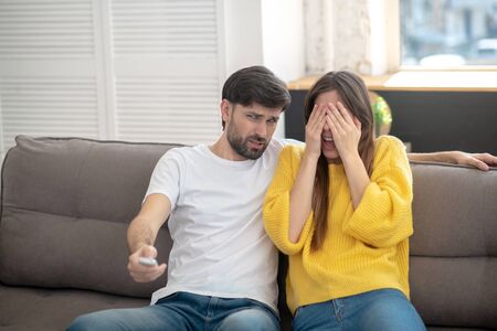 Dont want to watch. Bearded man with grimace and remote control in hand, girl in yellow sweater covering her face with her handsの写真素材