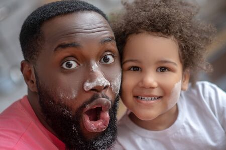 Having fun. Dark-skinned father opening his mouth and looking funny with face stained with flour, his daughter smilingの写真素材