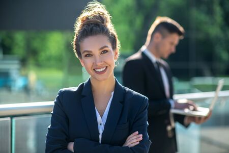 Successful and happy. Stylish successful businesswoman smiling standing clasped hands on chest, man with laptop behindの写真素材