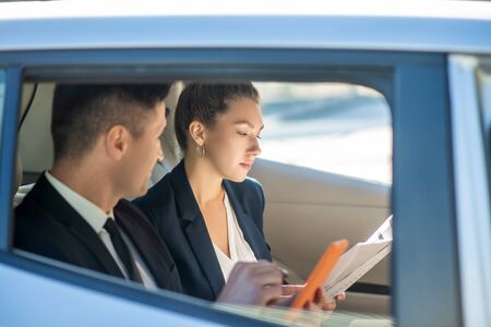 Success at work. Man in black suit and tie with tablet looking at woman carefully studying...