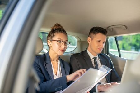 Busy business man with laptop and woman in glasses checking documents sitting in carの写真素材