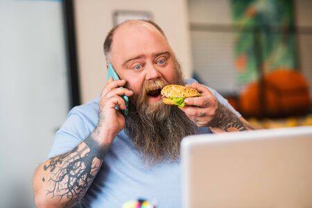 Eating at work. Bald man talking on the phone and eating a burgerの写真素材