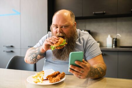 During lunch break. Bearded tattooed involved man eating appetite burger looking at smartphone in kitchen at tableの写真素材