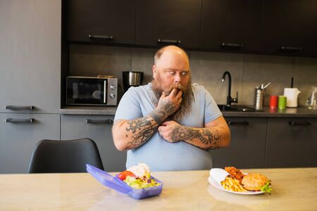 Desire to eat. Hungry big bearded man looking at burger and french fries on plate on kitchen table, hand near mouthの写真素材