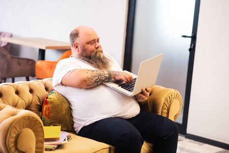Bald bearded plump man sitting with a laptop in hands and working onlineの写真素材