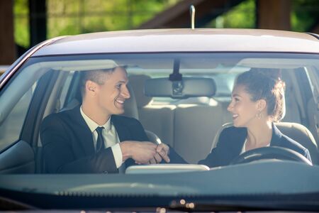 Young adult man and woman in business suits shaking hands in agreement sitting in a car, smiling