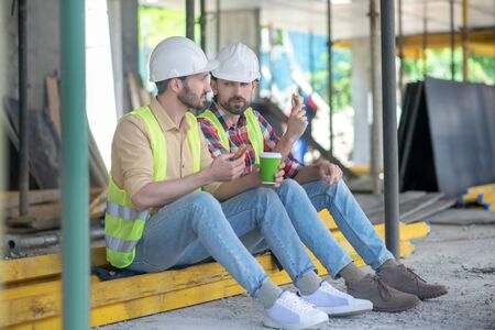 Building workers in yellow vests and helmets sitting on boards, having coffee with sandwiches,...