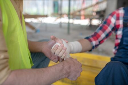 Building site. Close-up of building worker hands applying bandage on his coworker forearmの写真素材