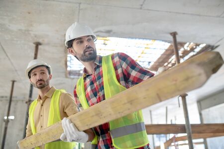 Building site. Building workers in yellow vests and gloves carrying heavy wooden board togetherの写真素材