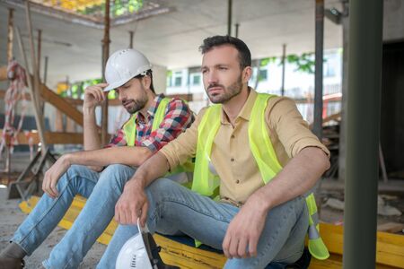 Building site. Tired building workers in yellow vests sitting on boards, one of men holding his helmet in his handの写真素材