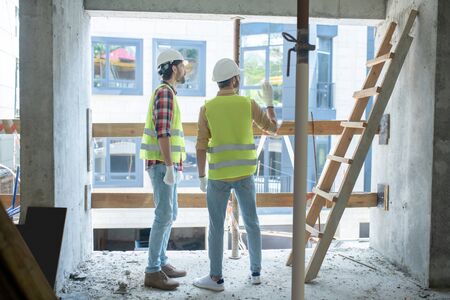 Building site. Building workers in yellow vests and gloves standing back to camera near the window,...