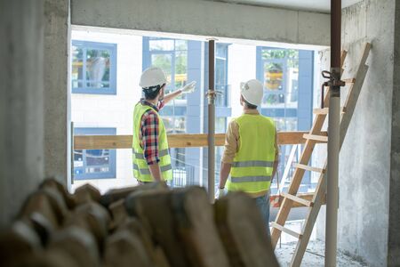 Building site. Building workers in yellow vests and gloves standing back to camera near the window,...