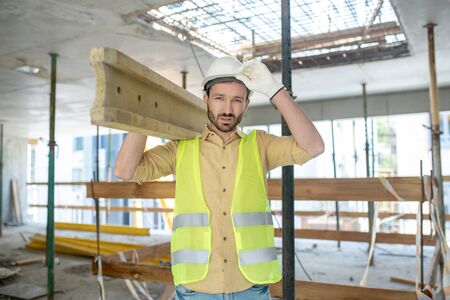 Building site. Tired building worker in yellow vest and gloves carrying wooden board on his shoulder, touching helmet with his handの写真素材