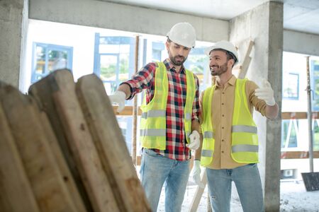 Building site. Building workers in yellow vests and gloves standing near pile of boards, discussing...