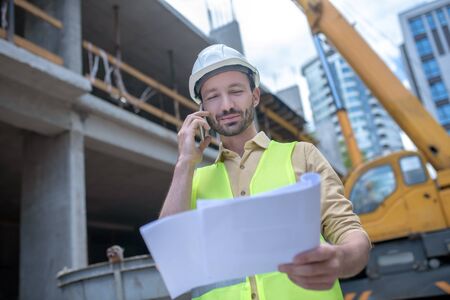 Building site. Building worker in helmet and yellow vest holding blueprint, talking on the phone