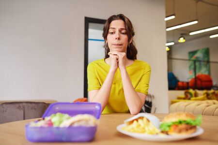Indecision, choice. Young indecisive woman with a grimace sitting at a table looking at a lunchbox and a plate of foodの写真素材